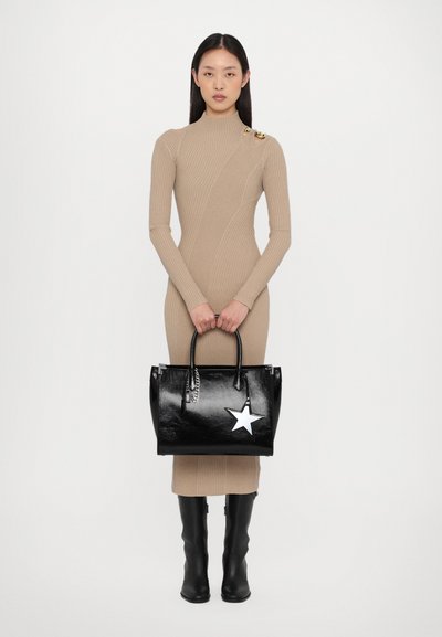 Sac en cuir noir en forme de sac fourre-tout avec un accent étoile blanche, de forme structurée et avec des anses doubles, associé à une robe en jersey beige ajustée et des bottes noires.