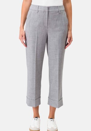 Femme portant un pantalon gris coupé avec des revers retroussés et des baskets blanches, debout devant un fond uni.