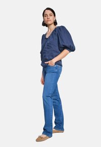 Navy blue puff-sleeve top with a scalloped neckline paired with light blue fitted jeans and beige flats, standing on a plain background.