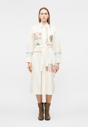 Γυναίκα που φοράει λευκό midi πουκάμισο-φόρεμα με πολύχρωμη γεωμετρική και ανθοκομική κεντητική, ζώνη δεμένη στη μέση και καφέ μποτάκια αστραγάλου, στέκεται μπροστά σε λευκό φόντο.