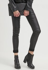 Pantalones de cuero sintético negros con acentos estriados y diseño ajustado. Llevados con un bolso de mano negro con tachuelas y botas de tobillo con tacón grueso.