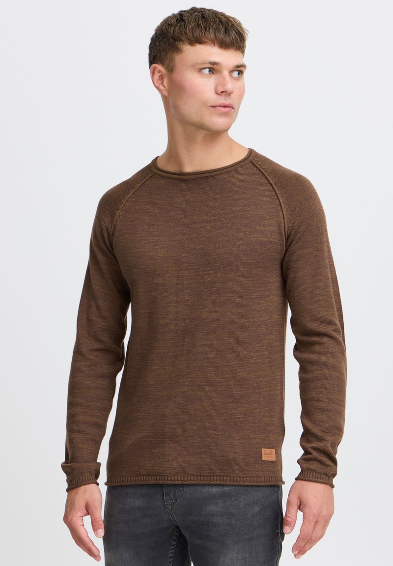 Pulover lung cu mânecă largă de culoare maro, cu un design texturat din tricot. Are un decolteu rotund și mansete cu zimți, precum și o etichetă mică cu logo la tiv.