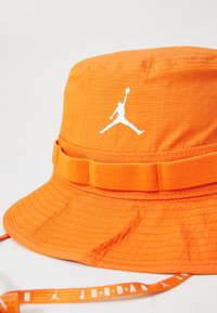 Gorra de cubo naranja hecha de tela texturizada con un ala ancha, que presenta un logo Jumpman blanco y una correa ajustable con marcas.