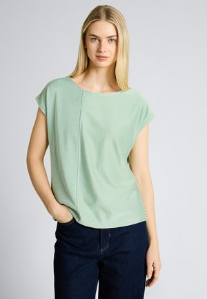 Femme blonde aux yeux bleus portant un haut vert clair à manches courtes et un jean foncé, debout avec une main dans la poche.