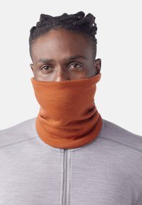 Un cache-cou orange porté par-dessus une chemise à manches longues grise. Le cache-cou est fait d'un tissu doux et texturé, avec un design tubulaire ajusté.