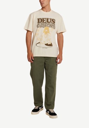 Camiseta de algodón beige con estampado gráfico en marrón, que presenta el texto "DEUS CUSTOMS" y un personaje. Combinada con pantalones de algodón verde oliva y zapatillas negras.