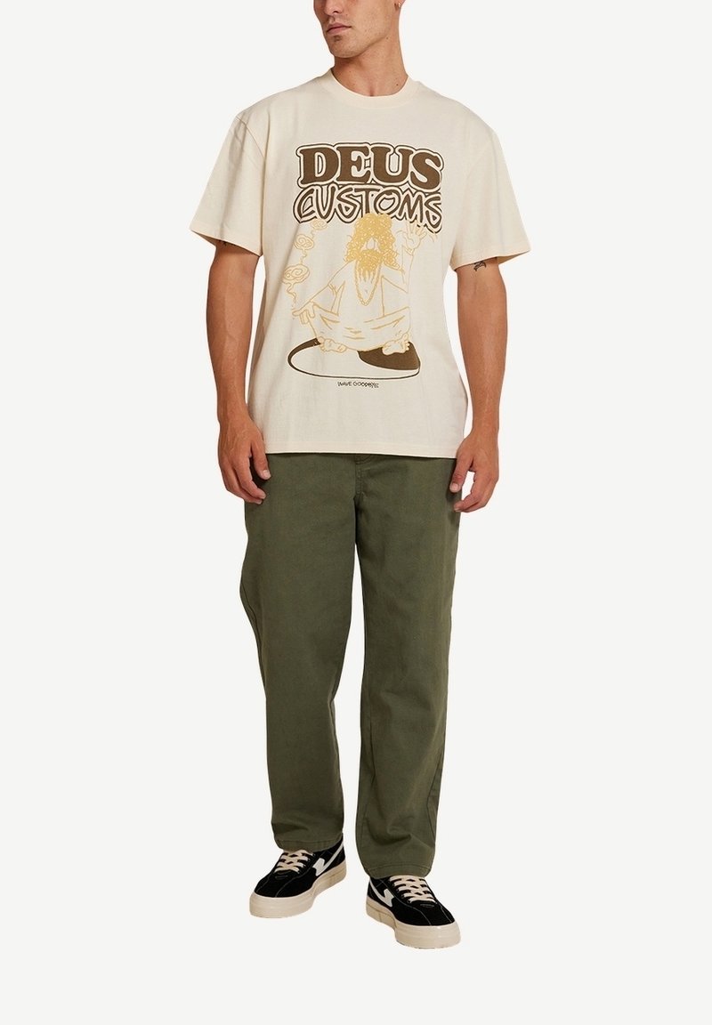 Camiseta de algodón beige con estampado gráfico en marrón, que presenta el texto "DEUS CUSTOMS" y un personaje. Combinada con pantalones de algodón verde oliva y zapatillas negras.