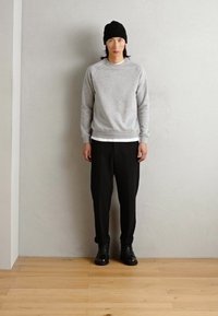 Pilkas megztinis su apvalia kaklo iškirpte, uždėtas ant baltos marškinėlių, derintas su laisvomis juodomis kelnėmis ir juodais šniūriniais batais, pabrėžiamas juodu skrybėle.