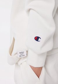 Vit sweatshirtärm med Champion-logotyp nära handleden och en hand i fickan, som visar ett tygetikett med texten "Var din egen mästare."