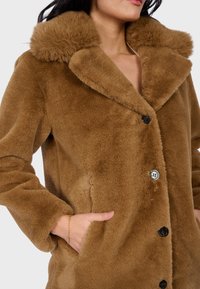Žena ve hnědém kožichu z umělé kožešiny s velkým límcem a zapínáním na knoflíky, ruka v boční kapse, na rukávu má tenký zlatý náramek.