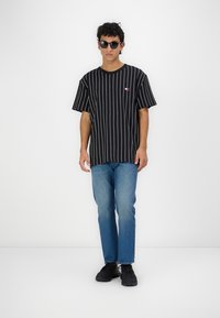 Svartstripete kortermet t-skjorte med en liten logo, kombinert med blå jeans og svarte joggesko. Modellen har solbriller på. Enkelt bakgrunn.
