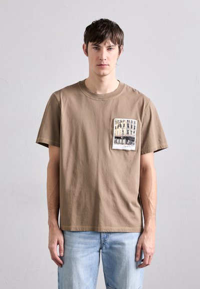 T-shirt en coton marron avec un col rond et des manches courtes. Présente une poche poitrine rectangulaire avec un impression photographique.