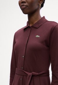 Rochie cu guler burgund, dintr-un material texturat, cu închidere cu nasturi în față și o curea legată în talie. Prezintă un mic logo verde și alb pe piept.