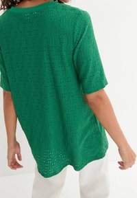 Top verde a maniche corte realizzato in tessuto tricotato testurizzato, con piccole aperture circolari, orlo arrotondato e vestibilità comoda.