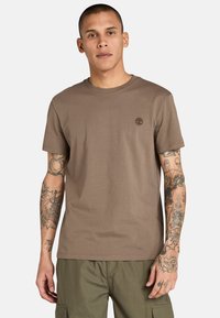 T-shirt in cotone marrone con scollo rotondo, maniche corte e un piccolo logo sul petto. Presenta tatuaggi visibili sulle braccia.