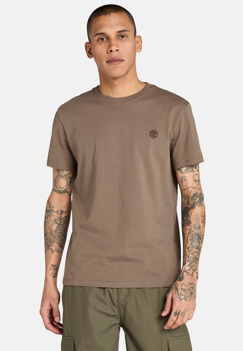 T-shirt in cotone marrone con scollo rotondo, maniche corte e un piccolo logo sul petto. Presenta tatuaggi visibili sulle braccia.