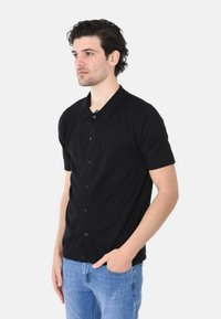 Camicetta nera a maniche corte con bottoni realizzata in un tessuto liscio, con colletto classico, bottoni sul davanti e un design aderente. Abbinata a jeans blu.