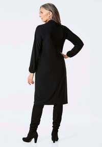 Schwarzes langärmliges Kleid mit taillierter Silhouette und Reißverschluss hinten, kombiniert mit kniehohen schwarzen Wildlederstiefeln. Glatte Textur und klassisches Design.