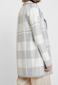 Frau in einem grau-weißen karierten Strickcardigan mit langen Ärmeln und Taschen, kombiniert mit hellbeigen weiten Hosen.