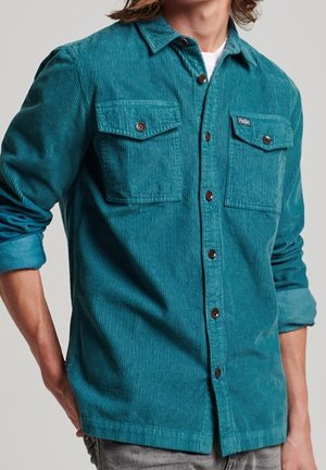 Mężczyzna w turkusowej koszuli sztruksowej zapinanej na guziki, z dwoma zapinanymi na guziki kieszeniami na piersi i podwiniętymi rękawami.