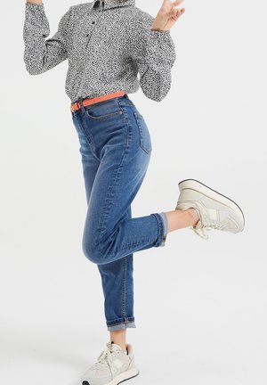Schwarz-weißer Polka-Dot-Hemd, hellblaue Denim-Jeans, orangefarbener Gürtel und beiges Sneakers mit strukturiertem Material und weißer Sohle.