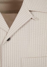 Beige gestreiftes Stoffhemd mit spitzem Kragen, schwarzem Knopf und aufgenähter Tasche im Brustbereich.