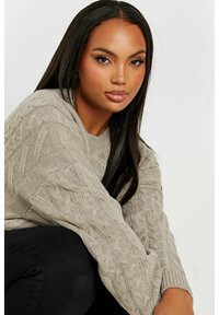 Jersey de punto grueso en gris claro con un patrón de trenzado texturizado, corte holgado y mangas anchas. Combinado con pantalones negros.