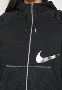 Svart zip-up hoodie med hög krage, silverfärgad Nike-logotyp på bröstet och texturerade axeldetaljer. Har metallisk dragkedja och snoddar.