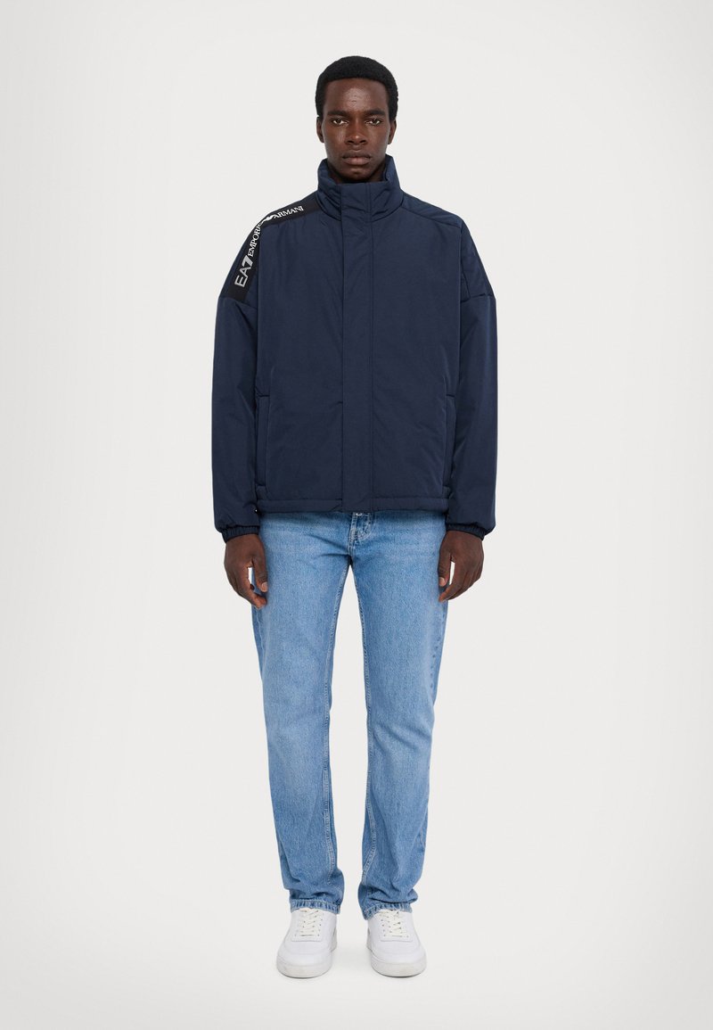 Chaqueta acolchada azul marino con cuello alto, cremallera frontal y logotipos en los hombros. Combinada con jeans de un tono azul claro y zapatillas blancas.