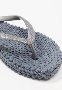 Grå flip-flop med en tekstureret vævet bund og glitrende sølvremme. Designet inkluderer en minimalistisk V-form på overdelen.