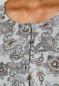 Tissu bleu clair présentant des motifs floraux et paisley complexes en noir et or. Encolure ronde avec des boutons décoratifs. Texture lisse.