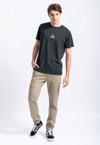 Zwarte katoenen t-shirt met een kleine kleurrijke afbeelding op de borst; gecombineerd met beige elastische tailleband broek en zwarte sneakers.