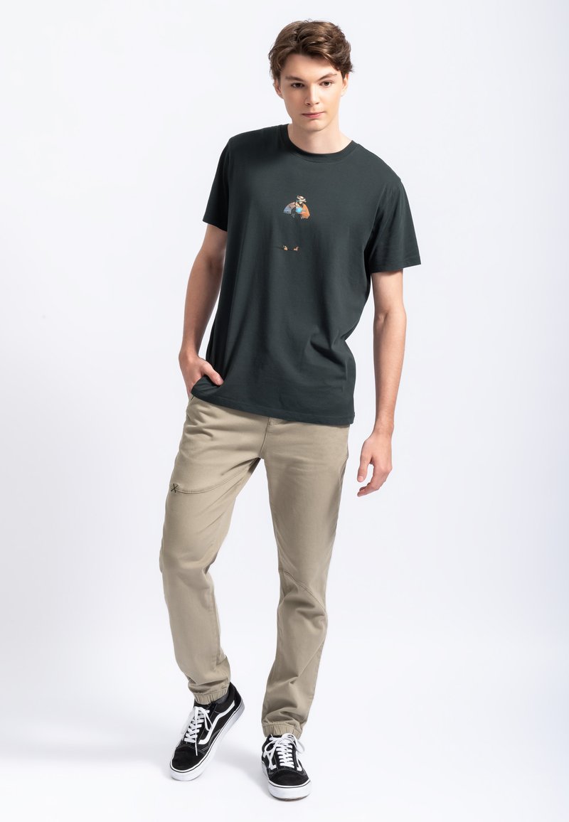 Zwarte katoenen t-shirt met een kleine kleurrijke afbeelding op de borst; gecombineerd met beige elastische tailleband broek en zwarte sneakers.