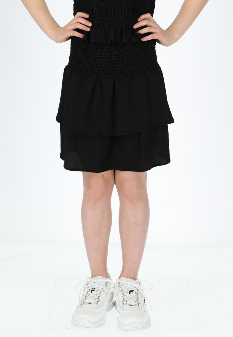 Robe noire à couches avec un corsage smocké et des accents en volants. Associée à des chaussures de sport blanches avec des détails gris et rouges.