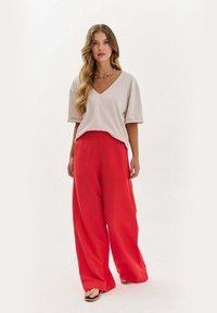 Žena s dlouhými vlnitými vlasy, oblečená v volném béžovém topu s výstřihem do V, širokých červených kalhotách a sandálech, stojící proti jednobarevnému světlému pozadí.