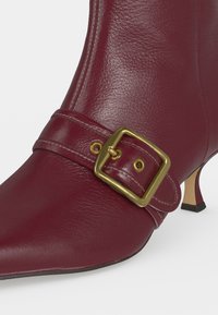 Gros plan d'une bottine en cuir rouge avec bout pointu, sangle à boucle carrée dorée et talon bas et fin.