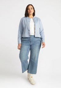 Chaqueta corta de color azul claro con cuello redondo, cierre de presión en el frente y bolsillos laterales, combinada con jeans de pierna ancha y zapatillas blancas.