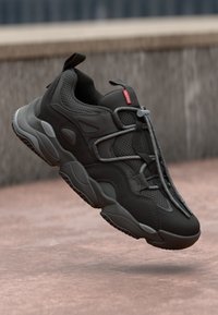 Black athletic shoe with mesh detailing, featuring elastic laces and a small red logo, positioned above a textured light surface.