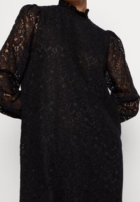 Vestido negro de encaje con largas mangas abullonadas, un cuello alto con borde rizado y un patrón floral de encaje en toda la tela.