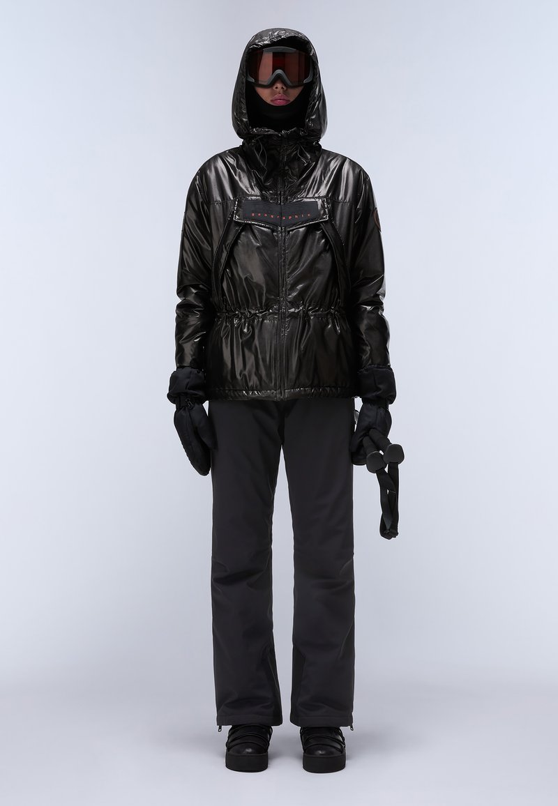 Chaqueta de esquí negra brillante con capucha, cintura con cordón y cremallera frontal. Combinada con pantalones de esquí negros y guantes. Los accesorios incluyen gafas de esquí.