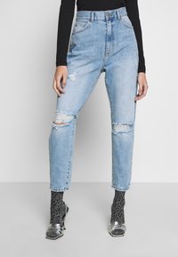 Kvinna klädd i ljusblå slitna jeans, silvriga öppna klackskor, svarta glittriga strumpor och en svart långärmad topp, stående mot en vit bakgrund.