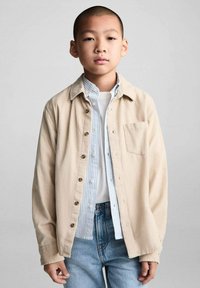 Camicia di corduroy beige con bottoni, indossata sopra una camicia a righe blu chiaro e bianca e una maglietta bianca. Accompagnata da jeans blu.