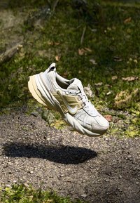 Chaussure de course légère en beige avec empeigne en mesh, accents texturés, et un grand swoosh doré. Dotée d'une semelle épaisse beige pour un meilleur amorti.
