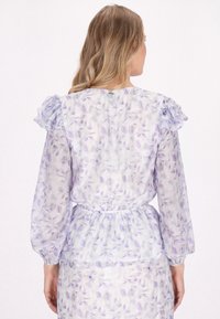 Letgå, gennemsigtig bluse med blomstermønster i lavendel og hvid, med flæser ved skuldrene og en sammensamlet talje. Lange ærmer inkluderet.