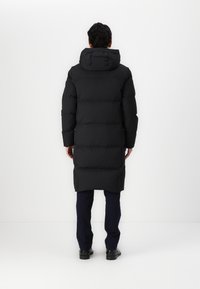 Man i en lång svart vadderad kappa med huva, marinblå byxor och svarta skor, som står vänd mot en enkel vit vägg.