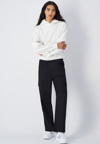 Sweat à capuche blanc avec une coupe décontractée, associé à un pantalon cargo noir avec des poches latérales, porté avec des baskets blanches.