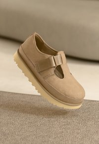 Beige suède schoen met uitsnijdingen, verstelbare band en dikke crèmekleurige rubberen zool. Zwevend boven een gestructureerd oppervlak.