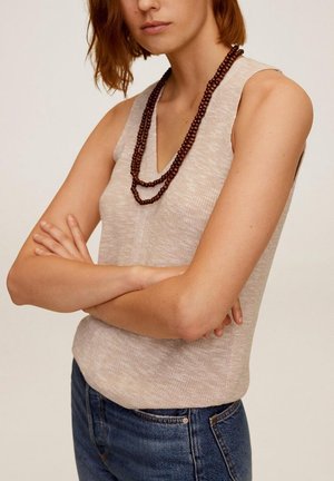 Frau mit kurzen roten Haaren trägt ein ärmelloses beiges Oberteil, eine mehrlagige dunkelbraune Perlenkette und blaue Jeans, die Arme verschränkt.