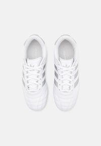 Des chaussures de sport blanches avec une tige en cuir texturé, trois rayures argentées de chaque côté et des lacets blancs. Comprend un col rembourré et un logo.