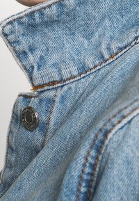 Modrá džínová bunda s vysokým límcem, kontrastním oranžovým prošitím a stříbrným knoflíkovým detailem na rameni. Textura vypadá měkce.